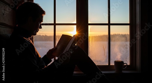 Silhouette of person reading book by window at sunset.