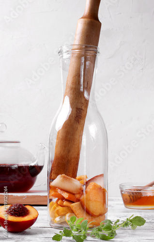 Nectarine and lemon pieces and a wooden muddler in a glass bottle on a white table. Arranged around it are black tea in a glass teapot, honey and mint sprigs. Making iced fruit tea. Step-by-step