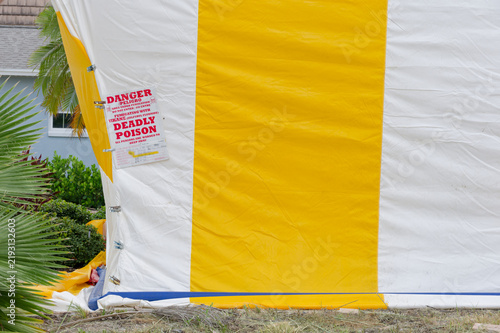 Bild auf Leinwand A yellow-and-white fumigation tent draped over a house behind a wooden fence, showing striped tarpaulin, metal fastenings and a red warning sign indicating poison, restriction and safety precautions