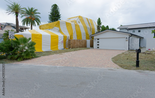 Canvastavla A yellow-and-white fumigation tent draped over a house behind a wooden fence, showing striped tarpaulin, metal fastenings and a red warning sign indicating poison, restriction and safety precautions