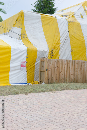 Фотография A yellow-and-white fumigation tent draped over a house behind a wooden fence, showing striped tarpaulin, metal fastenings and a red warning sign indicating poison, restriction and safety precautions
