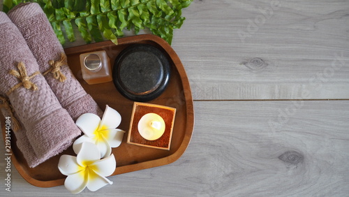 Принти на полотні Spa wellness arrangement set on a wooden tray, featuring rolled brown towels tied with twine, white plumeria flowers, a lit tealight candle in a wooden box, dark massage stones, flat lay top view