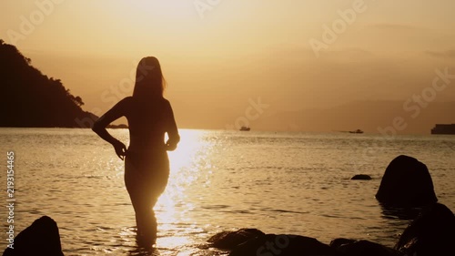 Slow motion backstage of a woman posing during photo session standing on the sea shore at sunset. Silhouette of a lady in swimwear posing and enjoying dusk at the tropical beach. Nature and fashion