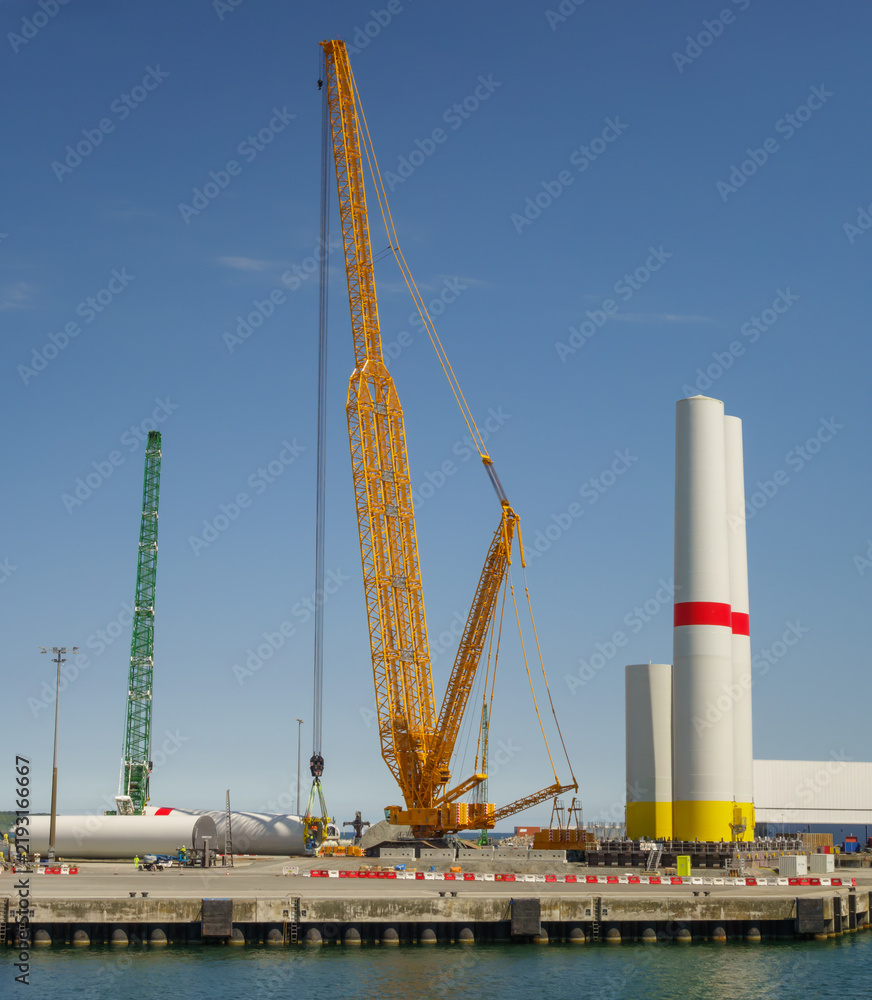 Fototapeta premium Giant Crawler Crane Assembling Wind Turbine Tower Sections at Port Harbor, Sustainable Offshore Renewable Energy Construction and Logistics