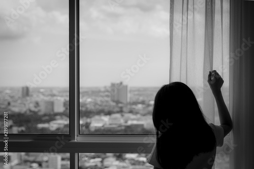 Obraz na plátne Silhouette of woman standing by large window overlooking urban cityscape, black and white scene expressing solitude, contemplation, isolation and quiet emotional reflection at home