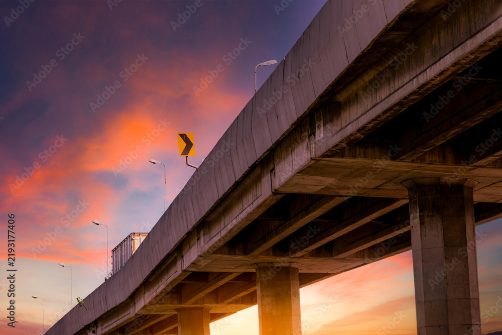 Obraz premium Elevated highway bridge against dramatic orange sunset sky with cargo truck, urban transportation infrastructure, road engineering and freight logistics concept.