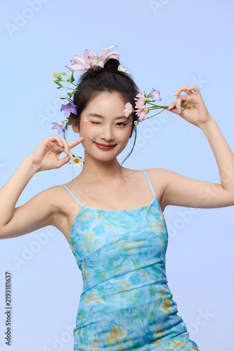 Картината върху платно Playful young woman with flowers in hair winking on pastel blue background