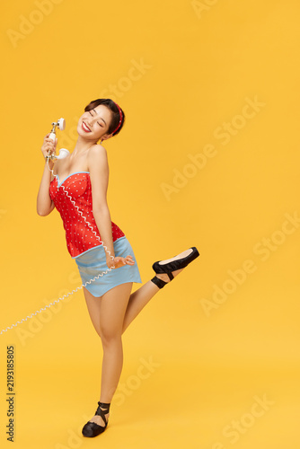 фотографія Playful woman in retro pin-up outfit holding vintage telephone on yellow backgro