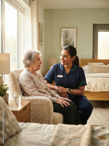 Elderly woman sitting in armchair talking with female healthcare worker in assisted living room