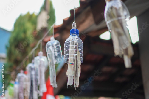 фотографія Clear plastic bottles repurposed as hanging wind chimes suspended from a wooden roofline, surrounded by greenery and soft natural light, evoking eco-friendly, rustic charm