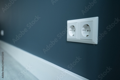A wall socket in the foreground with a freshly painted wall and white baseboard in the background, showing a renovated interior detail. 