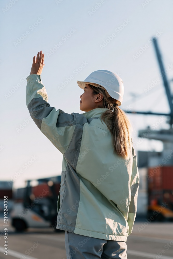 Fototapeta premium Female dock worker signaling at shipping port with safety helmet and jacket for logistics banner background and industrial transport design