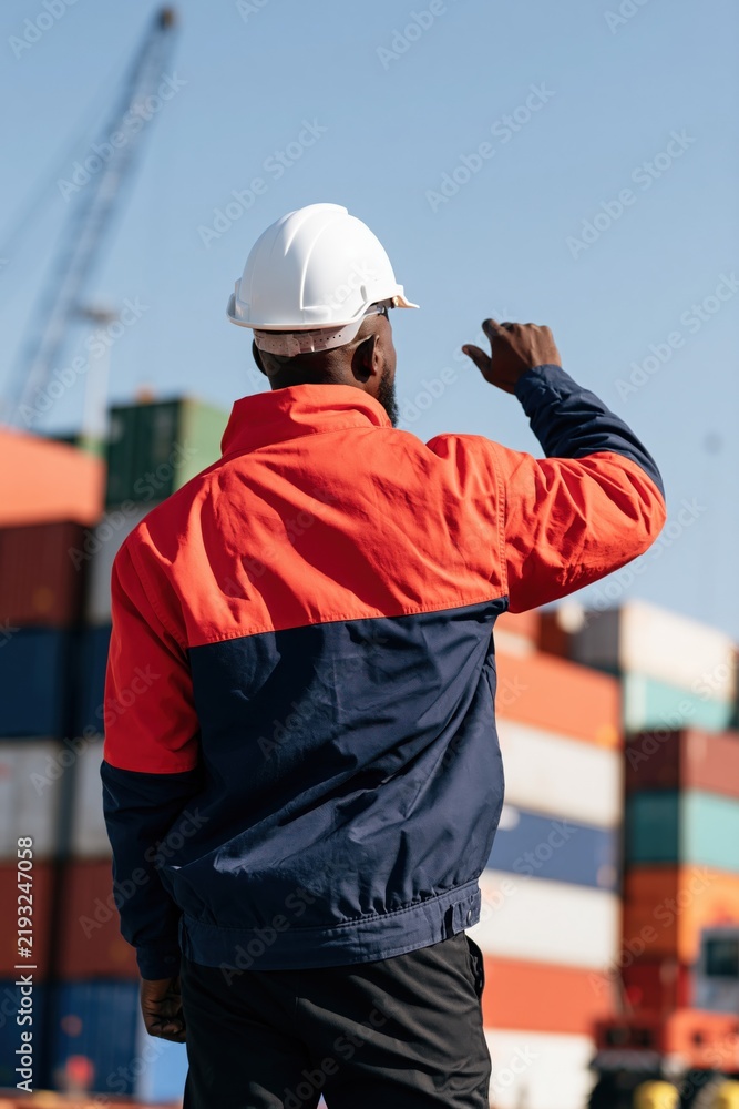 Fototapeta premium Male dock worker gesturing at shipping yard with safety helmet and high visibility jacket for industrial poster background and cargo handling design