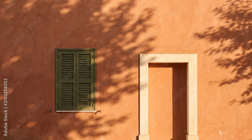 Obraz premium Sunlit mediterranean residential door with green shutter on warm terracotta wall under tree shadows