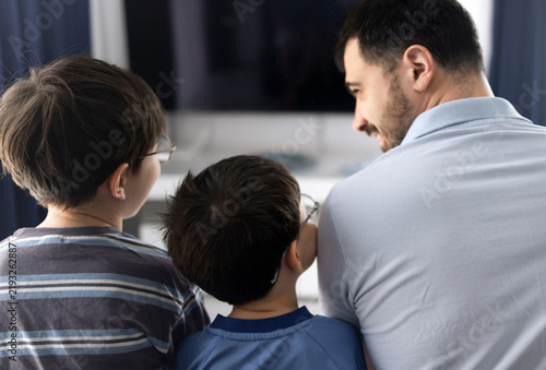 Father and his two sons sitting on a couch and watching television in a living room. The man holds a remote control while relaxing with the boys indoors