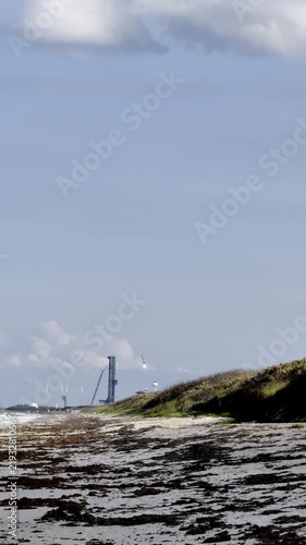 A rocket booster descends toward a coastal spaceport above a sandy beach and ocean shoreline. The landing burn, launch tower, dunes, clouds, and space coast landscape show a dramatic reusable rocket