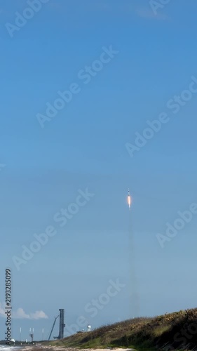 A rocket booster returns through a clear blue sky toward a coastal spaceport near the ocean. The landing burn, smoke trail, launch tower, beach, and dunes show a dramatic reusable rocket recovery