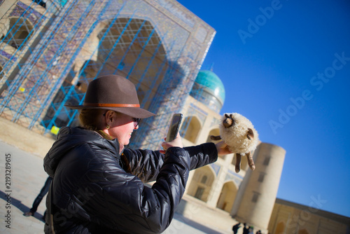 Cuadro en lienzo Tourist taking a photo of a plush souvenir sheep in front of the historic turquoise-tiled madrasa in Bukhara, Uzbekistan under a clear blue sky