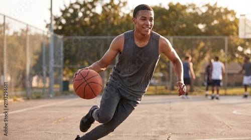 Full body action shot of smiling young man dribbling basketball on sunny outdoor court, active summer sports lifestyle concept.