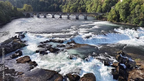Aerial view of Rhine Falls in Switzerland with powerful cascades rushing between rocky formations, a Swiss flag, cliffside viewing paths and a historic railway bridge.