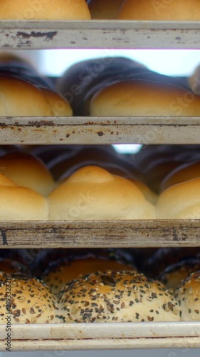rows of golden bread rolls rest on three worn metal shelves, close view with shallow depth of field, seeded tops and smooth plain tops, warm daylight. soft lit