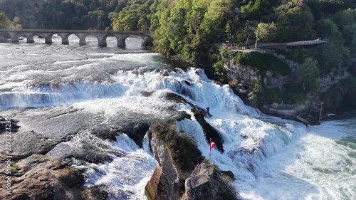 Aerial view of Rhine Falls in Switzerland with powerful cascades rushing between rocky formations, a Swiss flag, cliffside viewing paths and a historic railway bridge.