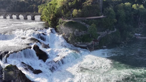 Aerial view of Rhine Falls in Switzerland with powerful cascades rushing between rocky formations, a Swiss flag, cliffside viewing paths and a historic railway bridge.