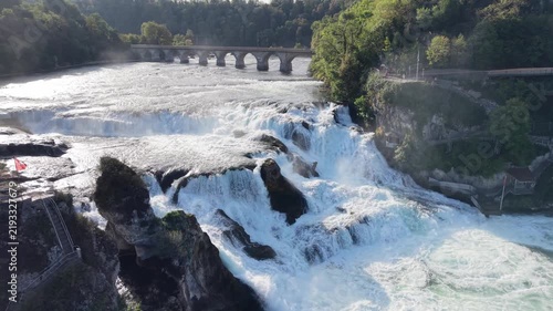 Aerial view of Rhine Falls in Switzerland with powerful cascades rushing between rocky formations, a Swiss flag, cliffside viewing paths and a historic railway bridge.