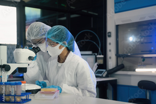 Two Scientists in Protective Gear Examining Samples Under Microscope in Modern Research Laboratory