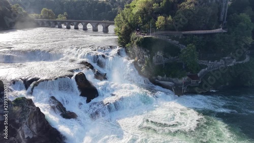 Aerial view of Rhine Falls in Switzerland with powerful cascading water, Schloss Laufen on the rocky hillside and a historic railway bridge surrounded by lush green forest.