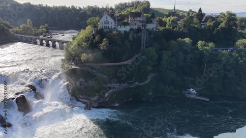 Aerial view of Rhine Falls in Switzerland with powerful cascading water, Schloss Laufen on the rocky hillside and a historic railway bridge surrounded by lush green forest.