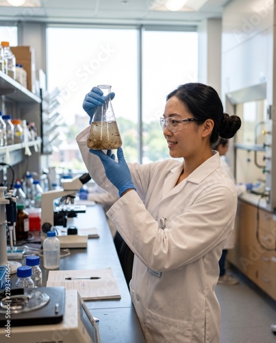 Young female Asian researcher in a white lab coat and safety goggles examining a liquid solution in a glass flask inside a modern scientific laboratory.