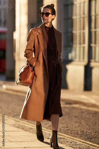 Fashionable woman in brown coat and sunglasses walks confidently on city street, showcasing stylish outfit against urban architecture in soft evening light