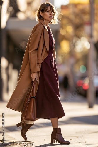 Fashionable woman in a brown coat and burgundy dress walks confidently on city street, holding a handbag with blurred urban background and warm sunlight