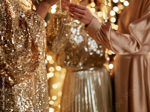 Three women in sparkling dresses toast with champagne glasses against a backdrop of warm bokeh lights, creating a festive and glamorous atmosphere for a celebration