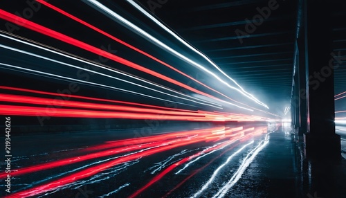 Long-exposure night photograph of vivid red and white vehicle light streaks flowing through a rain-soaked urban underpass, glossy pavement reflecting the glowing trails, dramatic perspective