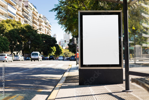 Vertical blank billboard at a bus stop shelter. Mockup lightbox with copy space for advertising or poster display in a city setting.