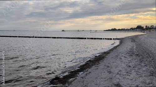 Calm Baltic Sea coast at sunset with wooden breakwater (Groyne), sandy shore, and pier under a cloudy sky