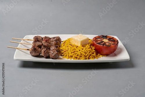 Chelo Kebab on white rectangular plate features grilled meat skewers, yellow rice, small block of butter, grilled tomato. Concept of grilled meat with saffron rice butter.
