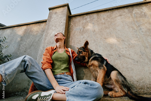 Fotografie Young woman sitting outdoors with a German Shepherd dog against a concrete wall, smiling and relaxing together in the yard during a sunny day with casual clothing