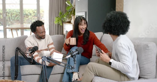 Diverse hosts podcasting with mics, left host opening white box, gesturing, holding mug on sofa