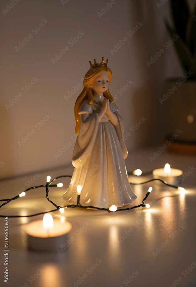 Fototapeta premium Spirituality and peace, ceramic angel figurine with prayer hands and crown surrounded by warm fairy lights and candles for holiday home decor