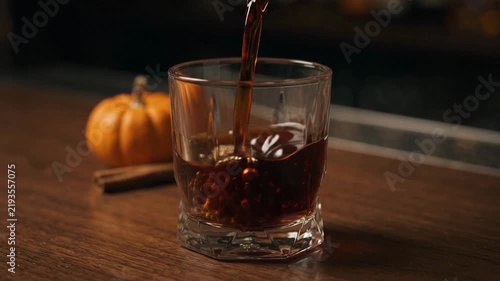 Elegant Halloween cocktail with rich dark liquid being poured into glass next to small pumpkin and cinnamon stick.