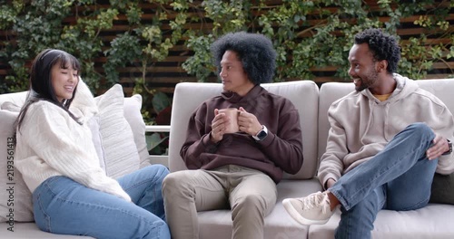 Diverse friends sitting on light outdoor sofa, center person picking up mug and sharing story