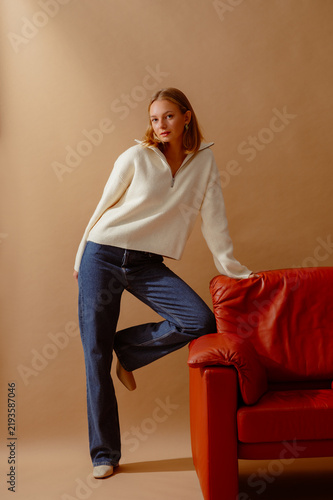Fashionable confident woman wearing trendy white knitted zip neck sweater, blue wide leg jeans, leather ankle boots, posing with brown armchair on beige backdrop. Full-length studio portrait
