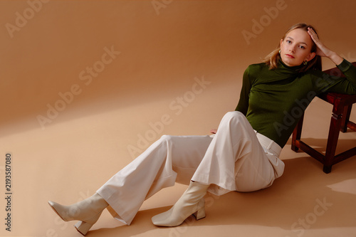 Fashionable confident woman wearing elegant olive green turtleneck top, white wide leg jeans, leather ankle boots, posing on beige backdrop. Full-length studio portrait. Copy, empty space for text