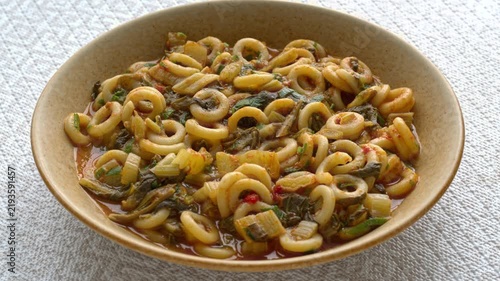 Authentic Italian Anelli Siciliani con Bietole e Pomodori dish. A brown ceramic plate with ring-shaped pasta, slowly rotating on a bright background with a beige geometric pattern.