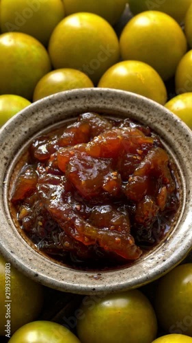 Macro shot of artisan green cherry tomato jam in a clay bowl. Slow, smooth rotation under soft light, with blurry fresh raw tomatoes used as jam ingredients filling the background. Vertical video.
