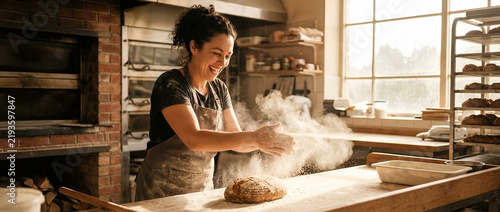 Young male baker with dark curly hair and medium brown skin tone dusting flour over fresh bread dough on wooden counter in rustic bakery kitchen with brick oven and natural light
