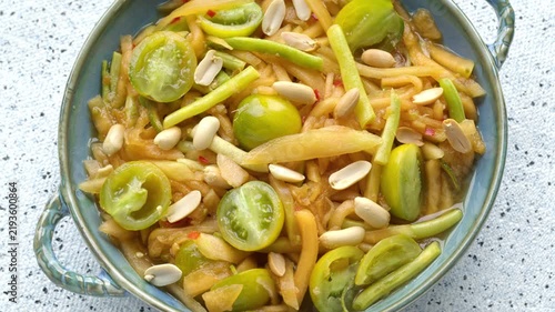 Top view of an alternative Thai som tam salad with stripped orange melon, yellow string beans, green grape cherry tomatoes, and roasted peanuts in a blue-green ceramic bowl. Table spin.